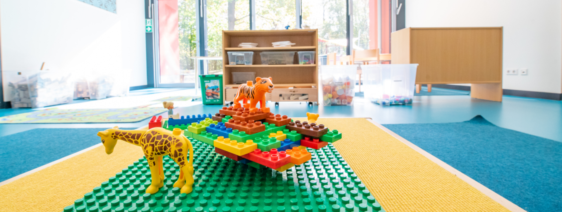 Im Vordergrund sind Lego-Duplo-Tiere und -Bausteine auf dem Boden, im Hintergrund ein Kita-Gruppenraum