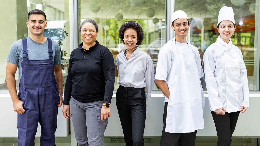 Ein Teamfoto mit verschiedenen Mitarbeitenden: einem Hausmeister, zwei Verwaltungsangestellten und zwei Mitarbeitenden aus der Gastronomie