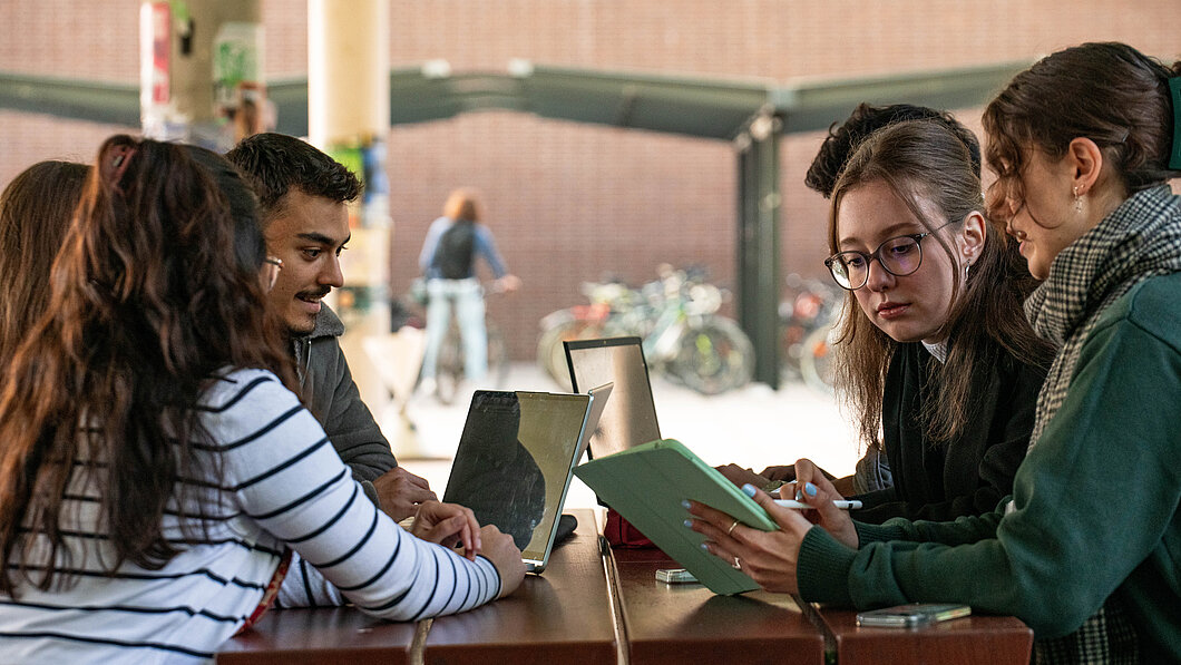 Foto von Studierenden im Gespräch an einem Tisch mit Tablet und Laptop