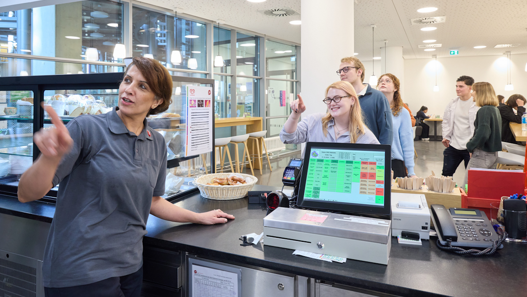 Kassiererin im Café Rabinstraße bedient eine Studentin; beide schauen auf die Angebotstafel an der Wand hinter der Kassiererin