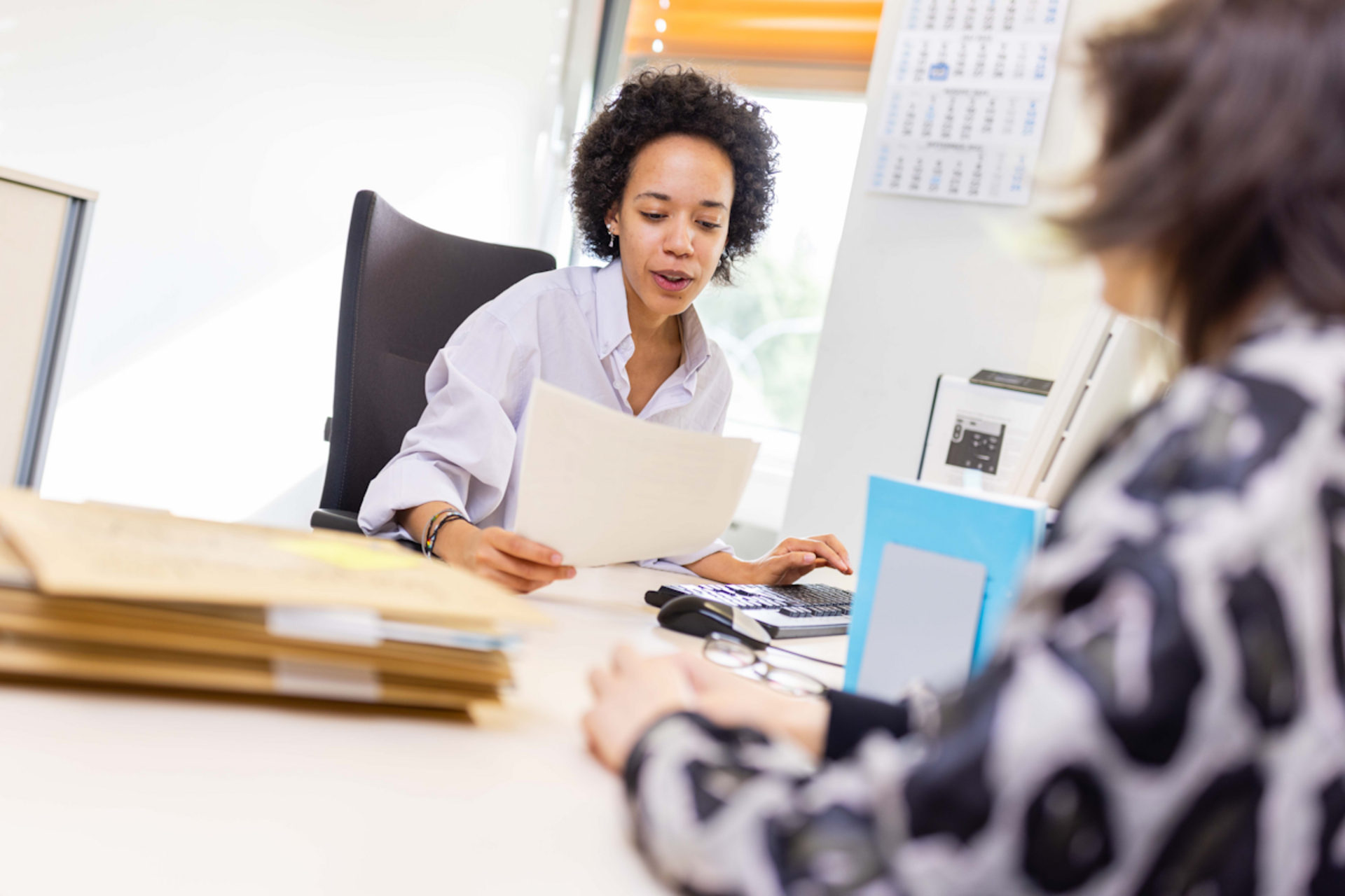 Frau sitzt an einem Schreibtisch. Sie hält ein Dokument in der Hand und spricht mit einer Frau, die ihr am Schreibtisch gegenüber sitzt. Auf dem Schreibtisch liegen Akten.