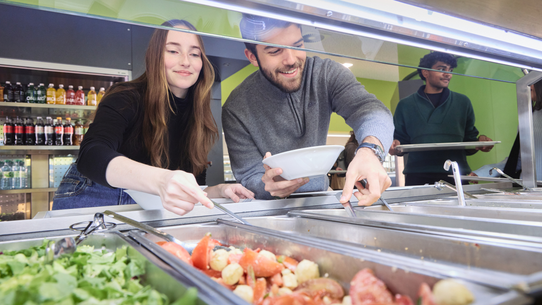 Zwei Studierende beugen sich über die Salatbar in der Mensa CAMPO und bedienen sich.