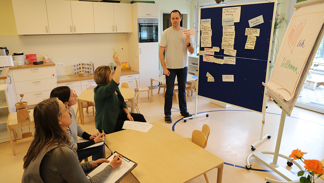 Bild von einer Team-Fortbildung, Mitarbeitende sitzen und eine Person steht am Pinbord