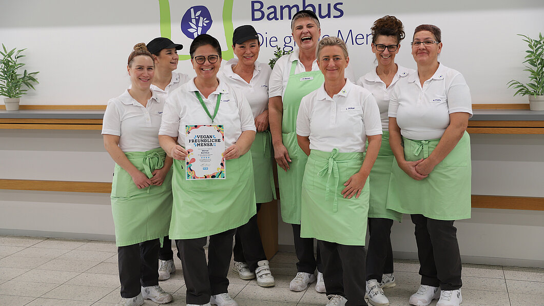 Foto vom Team der Mensa Bambus vor dem Logo der Mensa mit der PETA Urkunde