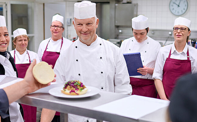 In einer Küche stehen Mitarbeitende um einen Tresen, auf dem ein Teller mit einem Burger steht.