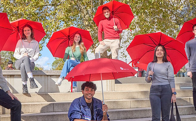 Eine Gruppe von neun Studierenden mit roten Regenschirmen vor der CAMPO