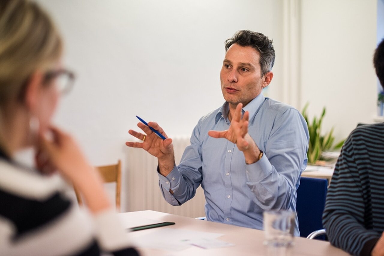 Ein Mitarbeiter in Führungsposition sitzt mit anderen Personen an einem Tisch und erklärt einen Sachverhalt.